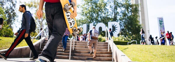 Student with skateboard walking up steps toward UCR logo silver metal scultpure near bell tower